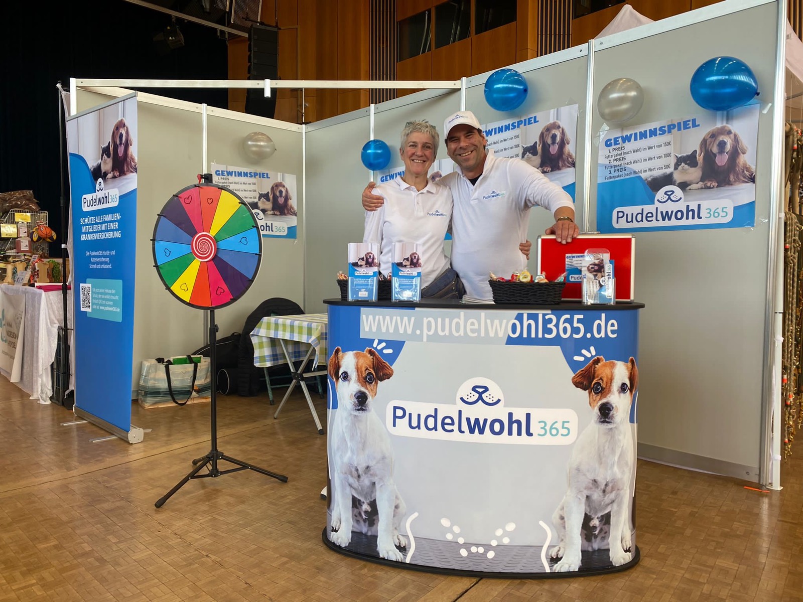 Zwei Mitarbeitende von Pudelwohl365 stehen lächelnd hinter dem Messestand mit Glücksrad, Roll-up und Gewinnspiel-Plakaten